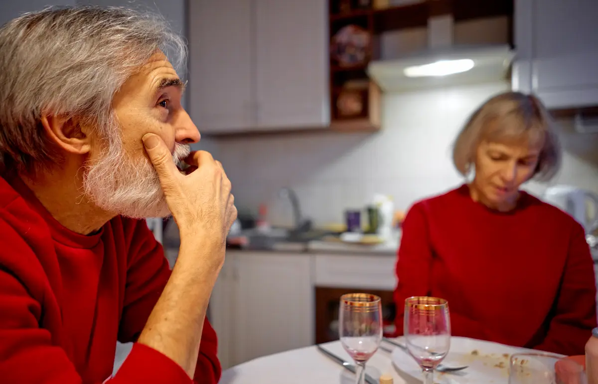 Older couple sit silently at a dinner table, with the man looking away thoughtfully while the woman looks down, suggesting emotional distance.